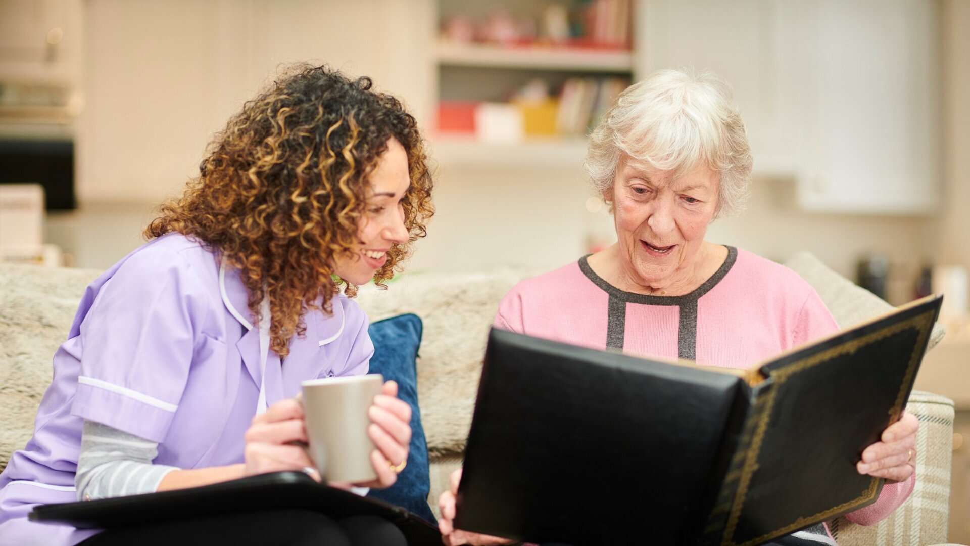 Female memory care resident sitting on a couch in her memory care apartment at Traditions at Lakeview Crossing with a caregiver