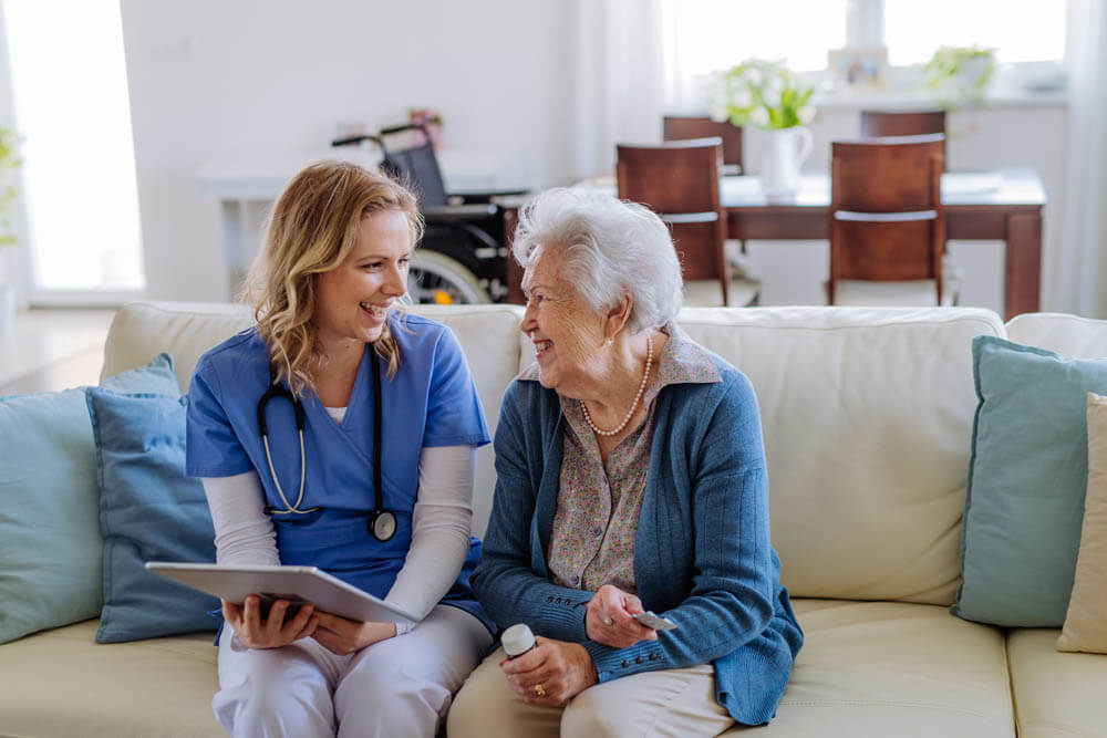Female nurse sitting on a sofa with a memory care patient, laughing and conversing, at Traditions at Lakeview Crossing in Groveport, OH.