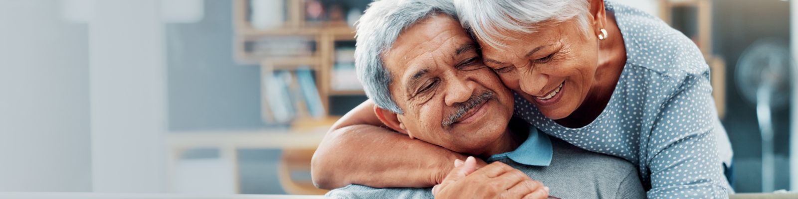 An older man with a mustache is sitting, smiling, an older woman hugging him from behind, leaning her head against his smiling