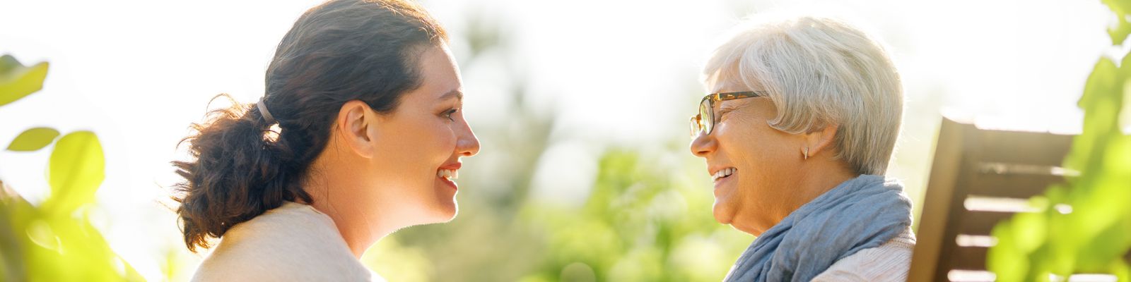 a young woman smiling affectionately at an older woman with glasses sitting in a sunlit area with foliage in the background