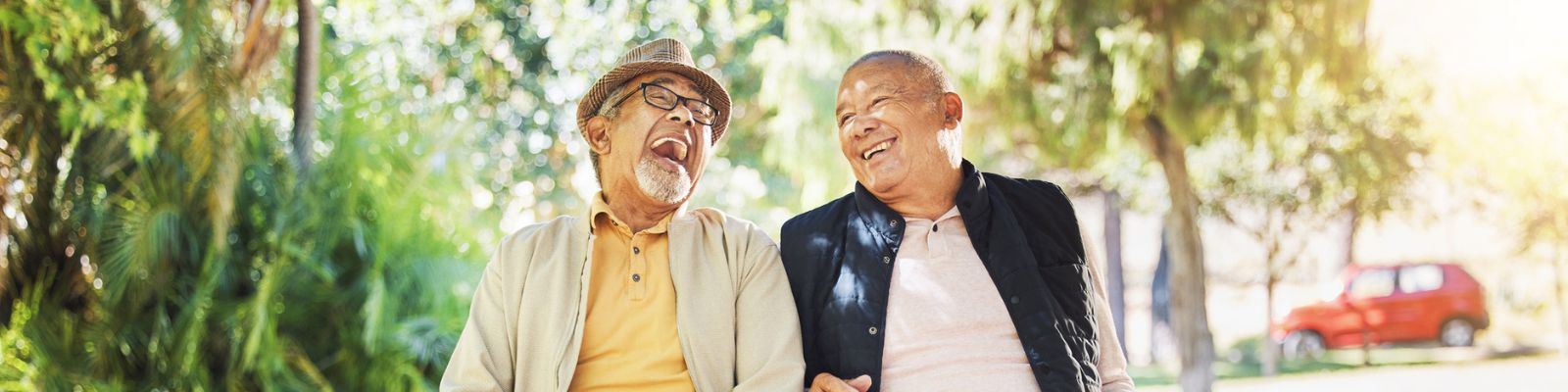 two elderly men dressed in layered clothing sitting outdoors in a park-like setting, smiling and laughing together
